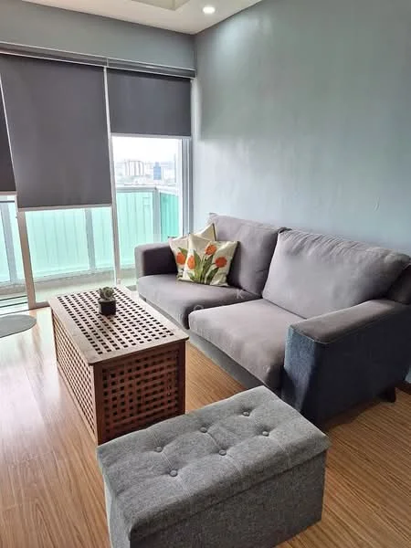 Living features: grey fabric 3-seater sofa, wooden coffee table with lattice design, grey upholstered ottoman, decorative throw pillow with floral pattern, laminate flooring, roller blinds on windows, floor-to-ceiling windows, green view from window, recessed ceiling lights, light blue painted walls; style: modern; view: city.