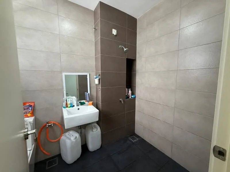 Bathroom features: grey tile walls, brown accent wall, black tile flooring, single-bowl ceramic sink, chrome faucet, mirror above sink, shower area with wall-mounted shower head, toilet, storage niche in shower, cleaning supplies visible, two plastic water containers, orange hose; style: modern.