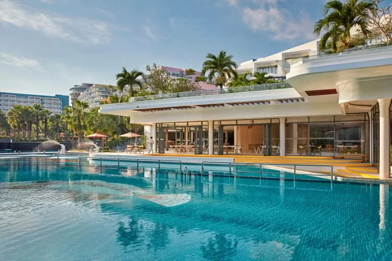 Exterior type: clubhouse; features: infinity pool, pool deck with loungers, surrounded by palm trees, large glass windows, outdoor dining area, tropical landscaping, sun umbrellas, modern architectural design; style: modern.