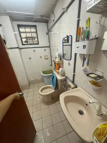 Bathroom features: white tiled walls, grey tiled flooring, single-sink vanity with white cabinet, chrome basin mixer faucet, framed mirror, one-piece toilet, blue toilet seat, shower area with handheld shower, wall-mounted storage shelf, toilet paper holder, soap dish, window for natural light; style: functional.