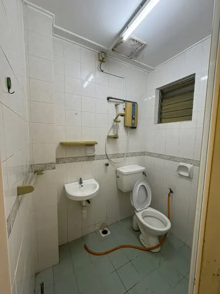 Bathroom features: white tiled walls, grey tiled flooring, single-bowl sink, chrome basin mixer faucet, one-piece toilet, shower with handheld showerhead, wall-mounted shower unit, window for ventilation, shelf above sink, toilet paper holder, drain in floor.
