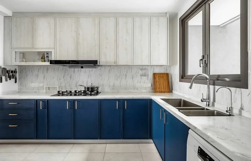 Kitchen features: blue cabinetry with gold handles, white marble countertop, white marble backsplash, stainless-steel double-bowl sink, chrome pull-down faucet, built-in gas cooktop, black range hood, wooden cutting board, tile flooring, large window with dark frames; style: modern.
