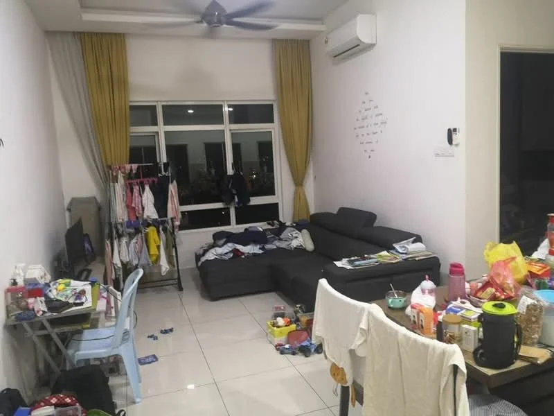 Living features: grey L-shape sectional sofa, flat-screen TV on stand, white painted walls, cream curtains, wall-mounted split AC, ceiling fan, white tile flooring, cluttered with various items, small dining table with blue chairs, clothes hanging on rack, scattered items on floor; view: city.