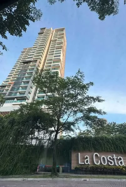 Exterior type: high-rise condominium; features: glass curtain wall, white painted facade, recessed balconies, landscaped entrance with greenery, signage with 'La Costa', multiple storeys, surrounded by trees; style: modern.