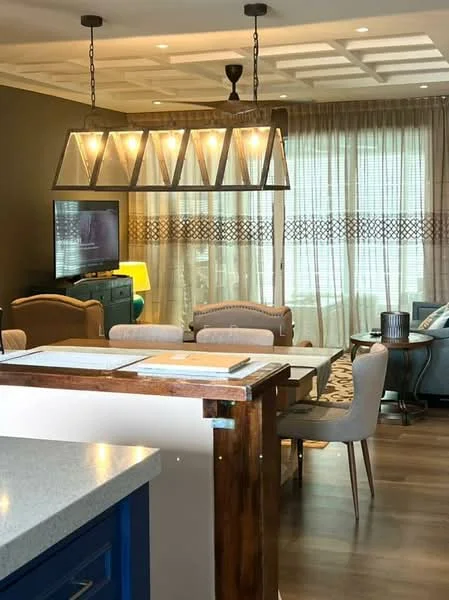 Dining features: wood dining table, upholstered dining chairs, chandelier with triangular shades, decorative wall paneling, large window with sheer curtains, TV mounted on wall, area rug with geometric pattern, wooden flooring, recessed ceiling lights; style: modern. Living features: fabric sofa, coffee table, side table, decorative lamp, wall-mounted TV, area rug, wooden flooring; style: modern. Kitchen features: granite countertop, blue cabinetry, stainless-steel appliances, open layout, pendant lighting; style: modern.