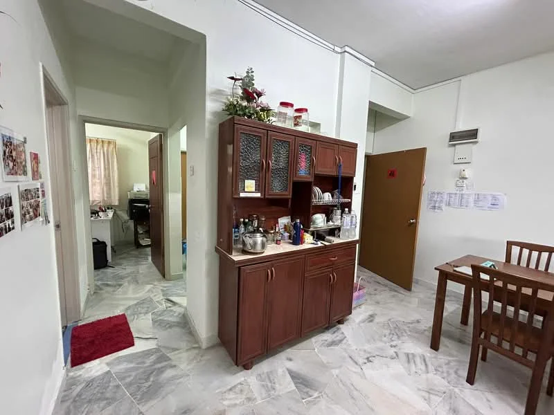 Kitchen features: brown wooden cabinetry, marble tile flooring, stainless-steel sink, chrome faucet, countertop with decorative items, open shelving, kitchen utensils, refrigerator visible in background. Dining features: wooden dining table, four wooden chairs. Room features: white painted walls, doorway leading to another room, light-colored curtains in background.
