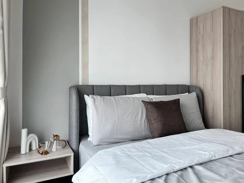 Bedroom features: queen-size bed, upholstered headboard in grey, white bedding, decorative brown throw pillow, nightstand with white finish, glass water carafe, ceramic vase, light-colored wooden wardrobe, white painted walls, curtains in light fabric; style: modern.