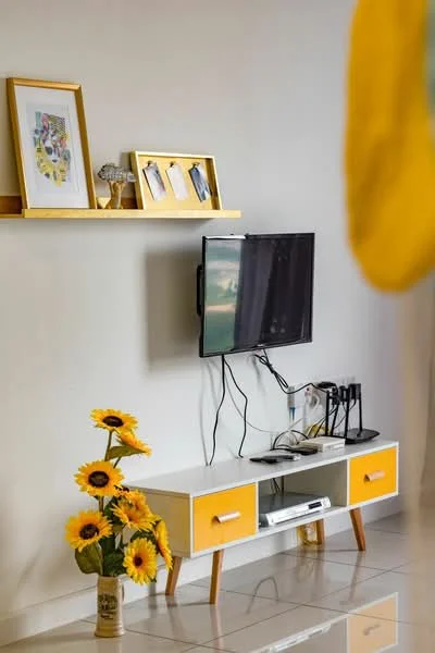 Living features: white walls, glossy tile flooring, wall-mounted TV, TV console with yellow drawers, decorative shelf with framed artwork, decorative frames, sunflower vase, electrical cables visible, modern design, natural light from window; style: modern.