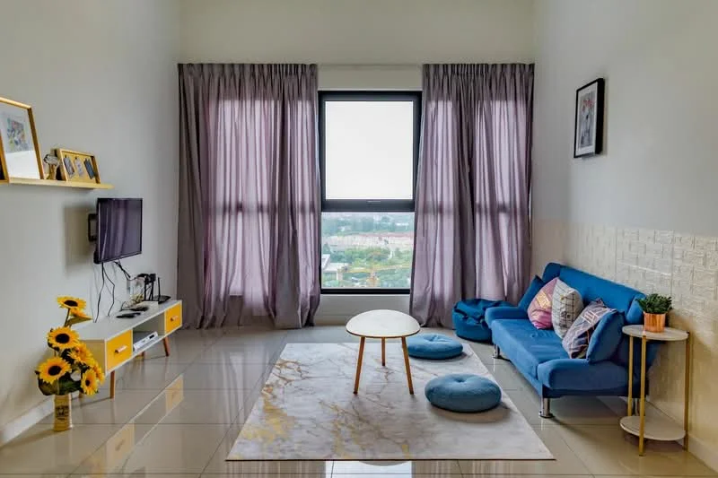 Living features: blue fabric sofa, round coffee table, two blue floor cushions, wall-mounted TV, shelf with decorative frames, large window with city view, purple sheer curtains, marble tile flooring, textured wall paneling, decorative houseplant, sunflower arrangement; style: modern; view: city.