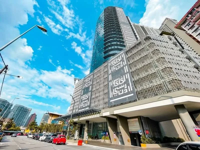 Exterior type: high-rise condominium; features: glass curtain wall, decorative facade with geometric patterns, multiple balconies, approximately 30-storey high-rise, covered parking area, driveway with interlocking pavers, surrounded by urban landscape, street lighting, clear blue sky, urban context with nearby buildings; style: modern.