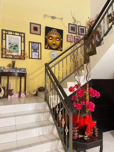 Room features: yellow painted walls, staircase with metal railing, white tiled flooring, decorative wall art, Buddha wall decor, wooden console table, vase with flowers, decorative plants, ornamental decorations, red floral arrangement, hanging decorative elements; style: traditional.