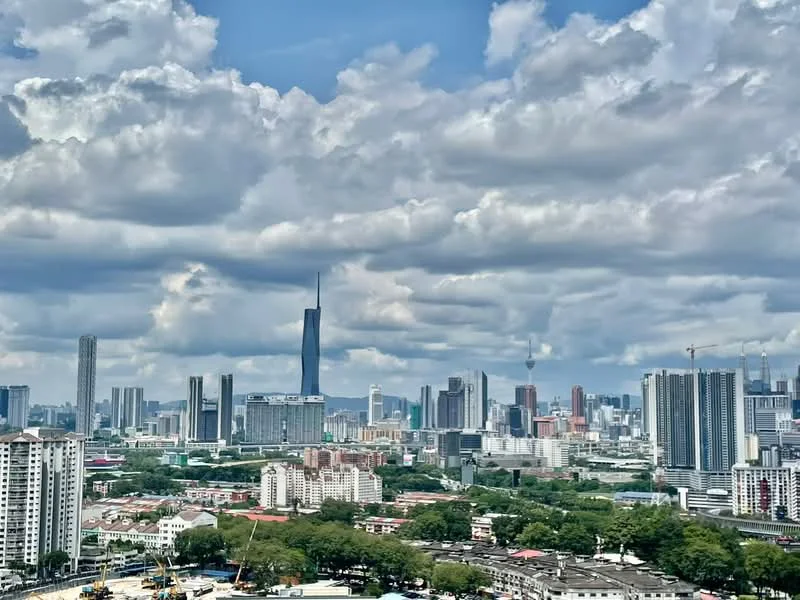 Exterior type: high-rise condominium; features: city skyline view, surrounded by greenery, urban landscape, various high-rise buildings, construction cranes visible, cloudy sky, Petronas Twin Towers visible, KL Tower visible; style: modern.