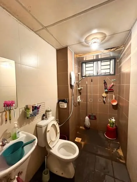 Bathroom features: one-piece toilet, shower area with handheld shower, wall-mounted shower head, mirror above sink, sink with faucet, tile flooring, brown tiled walls, ceiling light fixture, window for ventilation, storage rack with toiletries, cleaning supplies on floor.