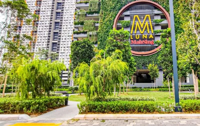 Exterior type: high-rise condominium; features: green wall with vertical gardens, large circular sign with 'LUNA' and 'MalaSing', manicured landscaping with shrubs, paved walkway, trees lining the pathway, modern architectural design, glass facade, surrounding greenery; style: modern.