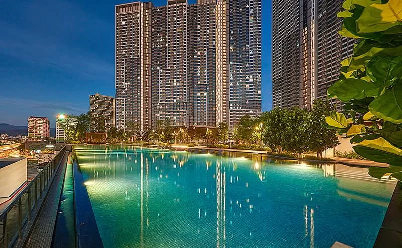 Exterior type: high-rise condominium; features: infinity pool, pool deck with loungers, surrounded by landscaped gardens, city skyline view, multiple high-rise buildings in background, night lighting, water feature, tropical landscaping; style: modern.