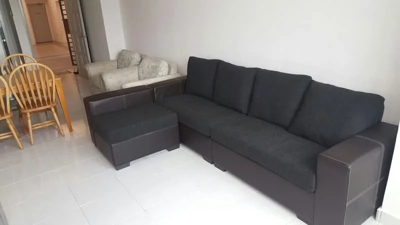 Living features: 3-seater fabric sofa in black, ottoman in dark brown, 2-seater fabric sofa in light color, wooden dining table with 4 chairs, white tiled flooring, plain white walls, natural light from adjacent room, open layout; style: modern.