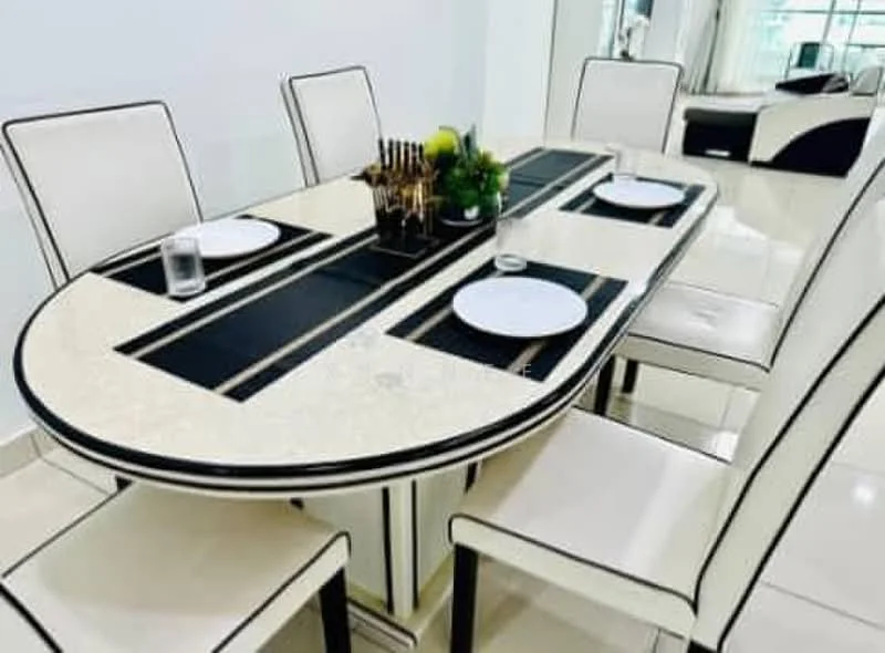 Dining features: oval dining table with marble top, white upholstered dining chairs with black trim, black placemats, glass water glasses, decorative plant in center, cutlery holder with utensils, polished tile flooring, white painted walls; style: modern.