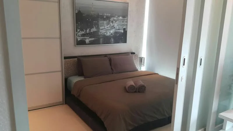 Bedroom features: king-size bed with brown bedding, upholstered headboard, two decorative pillows, two rolled towels, framed black-and-white artwork on wall, built-in wardrobe with sliding doors, soft beige carpet flooring, natural light from window, minimalist decor; style: modern.