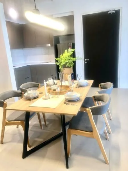 Dining features: wooden dining table, 4 dining chairs with grey upholstery, white plates, cutlery, decorative plant in vase, candles, light fixture above table, grey cabinetry in background, stainless-steel fridge visible, black door, light-colored flooring; style: modern. Kitchen features: grey cabinetry, white countertop, stainless-steel fridge; style: modern.