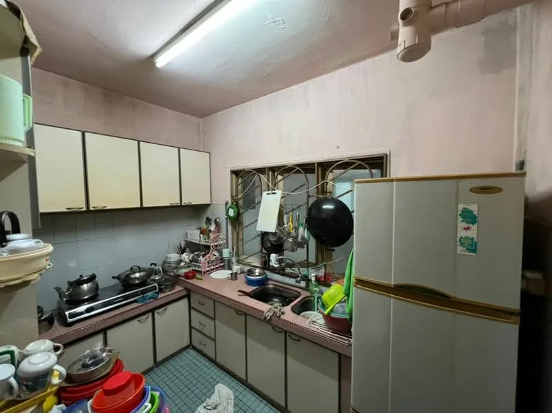 Kitchen features: pink painted walls, grey tiled flooring, L-shaped kitchen layout, double-bowl stainless-steel sink, chrome pull-down faucet, white cabinetry with brown trim, built-in gas stove, exhaust hood, refrigerator with top freezer, window with metal grilles, various kitchen utensils and pots, dish rack with dishes, countertop with cluttered items.