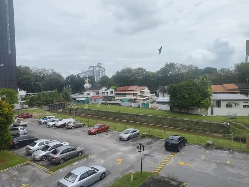 Exterior features: parking lot with multiple cars, greenery along the side, residential houses in the background, fenced area, cloudy sky, urban setting.