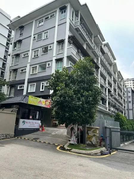 Exterior type: low-rise apartment block; features: grey painted facade, balconies with railings, multiple windows, entrance gate, landscaped area with trees, driveway with curved path, signage at entrance; style: modern.