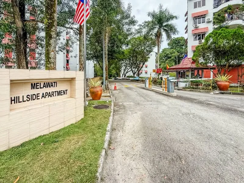 Exterior type: low-rise apartment block; features: entrance sign with 'Melawati Hillside Apartment', paved driveway, landscaped garden with plants, security gate, guard house at entrance, surrounded by trees, flagpole with Malaysian flag, parking area visible; style: modern.
