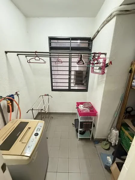 Laundry features: washing machine, grey tiled flooring, white painted walls, metal drying rack, clothes hangers on rod, window with security grille, plastic storage cart, mop, cleaning supplies, hose, shelf with storage boxes.