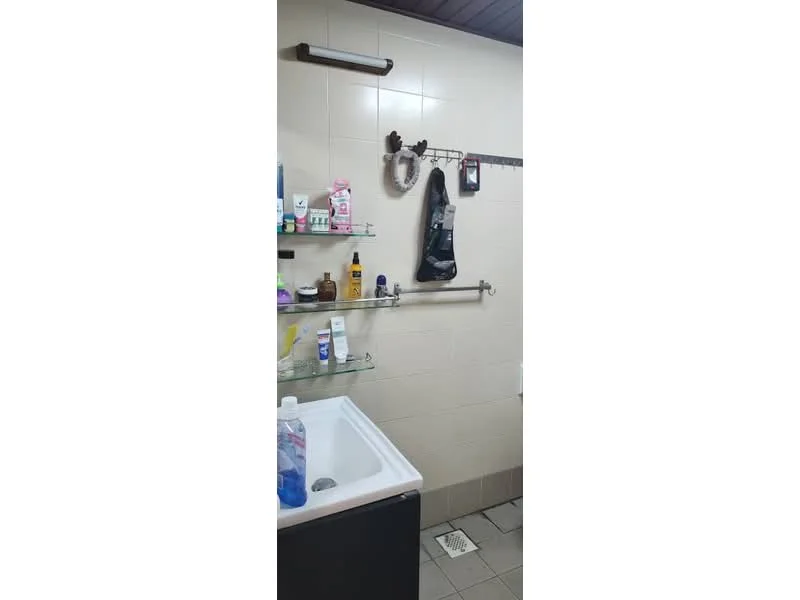 Bathroom features: white tiled walls, grey tiled flooring, glass shelf, metal towel rack, mirror, sink, various toiletries, shower area, shower curtain rod.