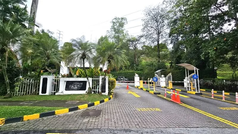 Exterior type: main entrance gate; features: gated entry, guard house at entrance, manicured landscaping, palm trees, stone-paved driveway, traffic cones, road markings, surrounded by greenery; style: modern.