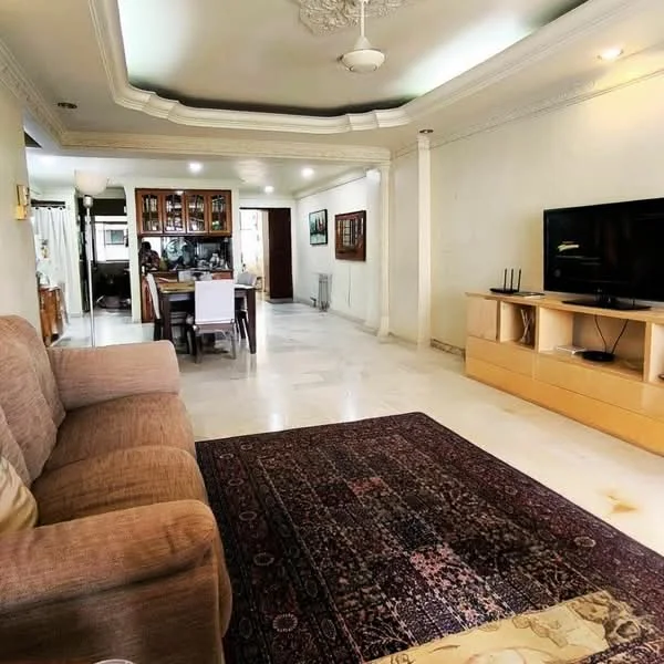 Living features: cream painted walls, marble flooring, tray ceiling with decorative molding, 5-blade ceiling fan, wall-mounted TV, wooden TV console, area rug with intricate pattern, sofa in beige fabric, dining area visible in background, glass-front cabinets, dining table with chairs; style: traditional.