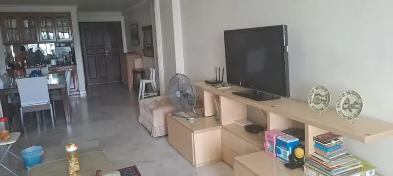 Living features: marble flooring, cream painted walls, wall-mounted TV, wooden TV console, 3-seater fabric sofa in beige, coffee table, ceiling fan, decorative plates on shelf, books on shelf, open shelving unit, dining area visible in background, doorway leading to other rooms; style: modern.