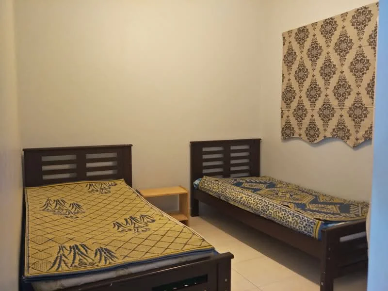 Bedroom features: two single beds with wooden frames, mattresses on both beds, decorative bedspreads with patterns, wooden bedside table, plain walls in light color, tiled flooring, window with patterned curtain.