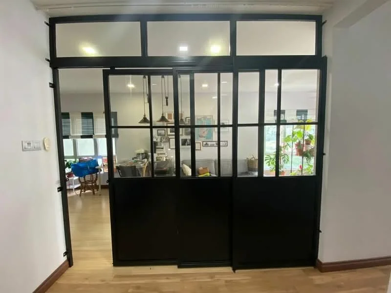Room features: black framed glass doors, wooden flooring, recessed downlights, wall-mounted light switch, white painted walls, visible living area beyond doors, decorative plants visible through glass, open layout; style: modern; view: living area.