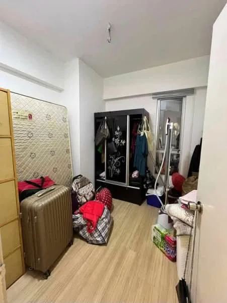 Room features: light wood flooring, white painted walls, built-in wardrobe with mirrored doors, queen-size mattress on bed frame, various bags and luggage, clothing hanging on hooks, window with sheer curtains, ceiling light fixture.