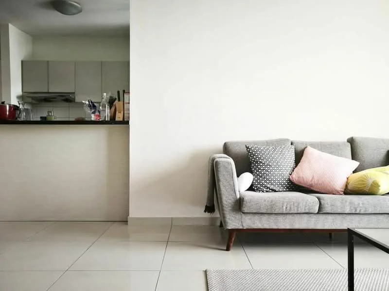 Living features: grey fabric sofa with wooden legs, decorative cushions in various colors, white walls, glossy tile flooring, open kitchen layout visible in background, kitchen cabinets in light color, kitchen countertop, recessed ceiling lights; style: modern.