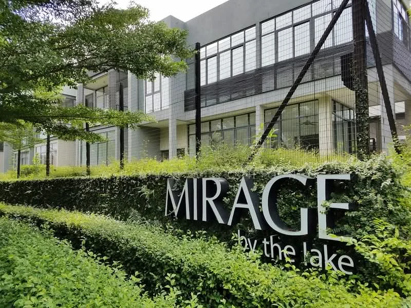 Exterior type: low-rise apartment block; features: modern facade with large windows, green landscaping with shrubs, signage with 'MIRAGE by the lake', fencing around the property, concrete structure, surrounded by greenery; style: modern.
