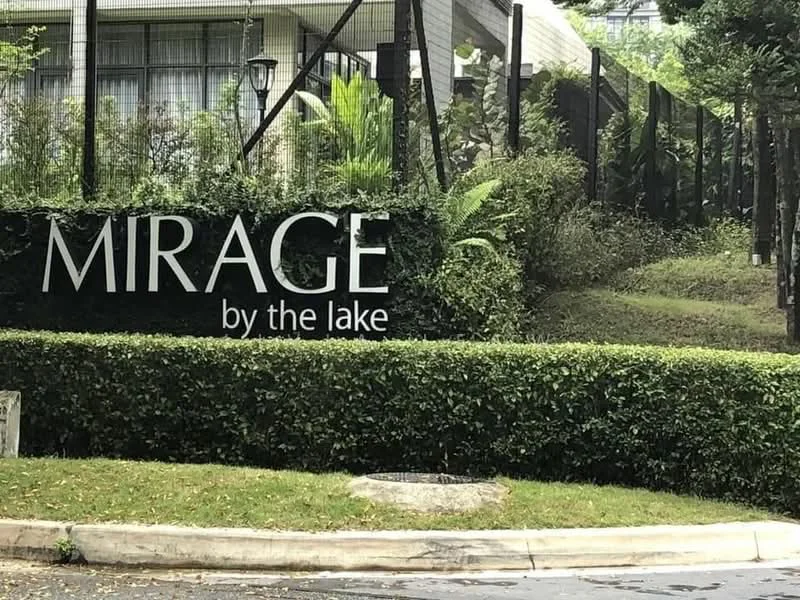 Exterior features: landscaped garden with hedges, signage with the name 'MIRAGE by the lake', fencing around the property, mature greenery, paved road, lawn area; style: modern.