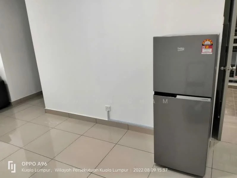Room features: grey fridge, white walls, tile flooring, power outlet on wall, neutral color scheme, corner of room visible, doorway leading to another room.
