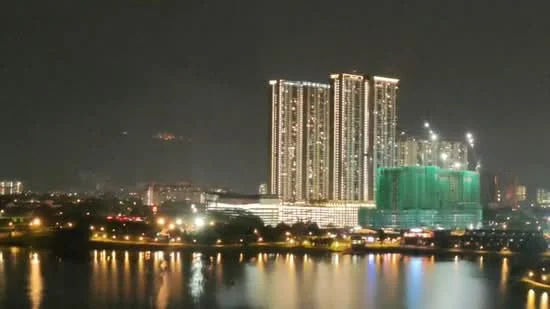 Exterior type: high-rise condominium; features: illuminated facade, glass curtain wall, multiple towers, waterfront view, surrounding urban landscape, night lighting, construction site with scaffolding, reflections on water; style: modern.