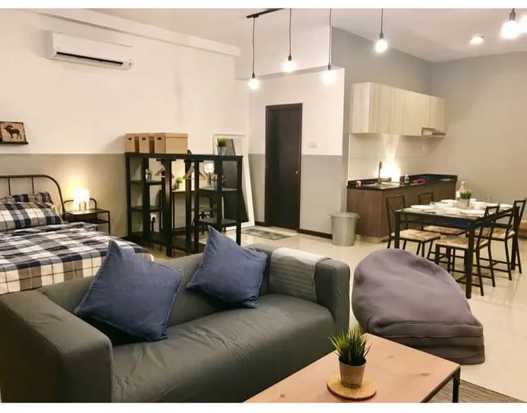 Living features: 3-seater fabric sofa in grey, 2 blue throw pillows, wooden coffee table, small potted plant on coffee table, bean bag chair, wall-mounted split AC, recessed downlights, floor tiles in cream, open shelving unit, framed artwork on wall, plaid bedspread on bed, queen-size bed, dining table with 4 chairs, kitchen cabinetry in light wood, black countertop, kitchen appliances visible; style: modern.