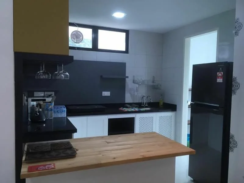 Kitchen features: black granite countertop, white cabinetry with mesh doors, black backsplash, stainless-steel fridge, built-in sink, kitchen island with wooden top, glass pendant lighting, wall-mounted shelves, kitchen utensils on countertop, water bottles on countertop, floor-to-ceiling window, white tiled walls; style: modern.