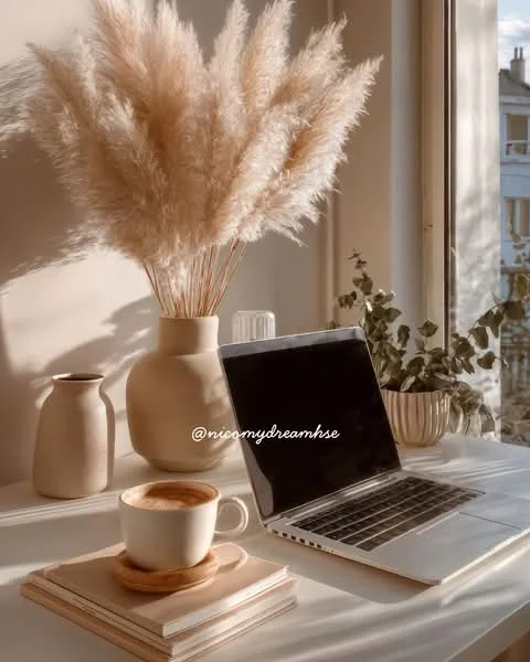 Room features: large window with natural light, wooden table, laptop on table, ceramic coffee cup, stack of books, tall vase with dried pampas grass, medium-sized vase, small potted plant, neutral color palette, minimalist decor; style: minimalist.