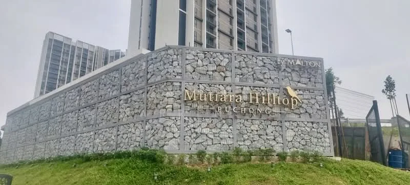 Exterior type: high-rise condominium; features: stone cladding on facade, prominent signage with 'Mutiara Hilltop', landscaped green area, surrounding grass, modern architectural design, multiple storeys visible, entrance gate, surrounding greenery; style: modern.