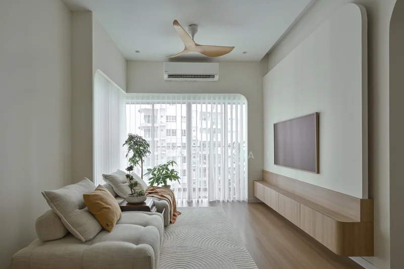 Living features: 3-seater fabric sofa in light beige, decorative cushions in orange and cream, wooden TV console, wall-mounted flat-screen TV, ceiling fan with 3 blades, floor-to-ceiling windows, sheer curtains, wooden flooring, indoor plant in ceramic pot, area rug with circular pattern, light-colored wall with curved edges, air conditioning unit mounted on wall; style: modern; view: city.