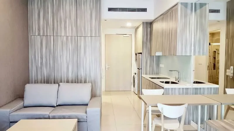 Living features: grey fabric sofa, coffee table, wall-mounted cabinetry, light-coloured tiled flooring, recessed ceiling lights, open-plan layout, neutral wall colour, kitchenette with countertop; style: modern. Kitchen features: white quartz countertop, double-bowl stainless-steel sink, chrome pull-down faucet, built-in oven, stainless-steel refrigerator, 2-burner electric hob, white cabinetry, washing machine; style: modern.