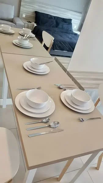 Dining features: light wood dining table, white ceramic dinner plates, white ceramic bowls, stainless-steel cutlery, white ceramic cups, minimalist table setting, neutral wall color, bedroom visible in background; style: modern.