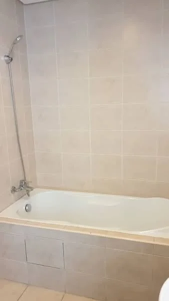 Bathroom features: built-in bathtub, chrome handheld shower, tile cladding, light beige wall tiles, white bathtub, single-lever faucet, tile flooring, shower head, storage cabinet under bathtub; style: modern.
