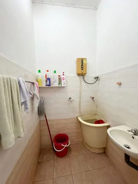 Bathroom features: white tiled walls, beige tiled flooring, single sink, wall-mounted faucet, toilet, shower head, water heater, cleaning supplies on shelf, mop, red bucket, towels hanging, soap dispenser.