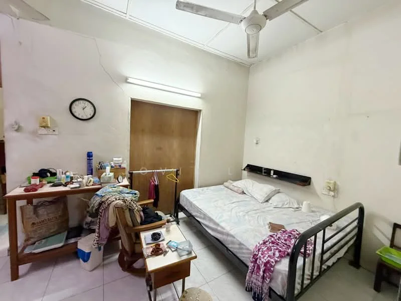Bedroom features: double bed with white bedding, wooden bed frame, nightstand with drawer, table lamp, wooden table with various items, clothes hanging on a rack, wall clock, ceiling fan, white painted walls, tile flooring, window with no visible treatment.
